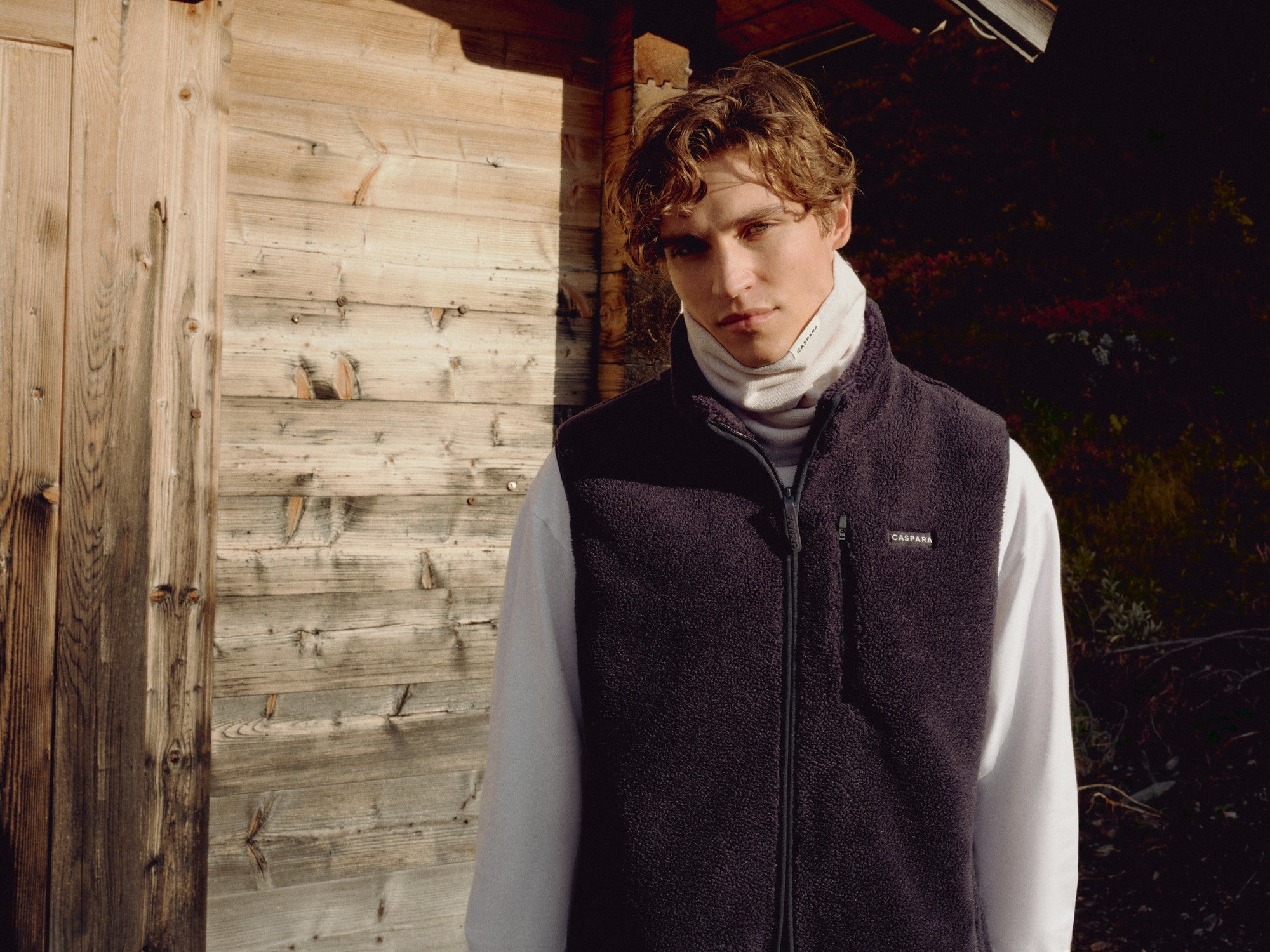 Man wearing a creme CASPARA Classic Neckwarmer standing by a wooden cabin.